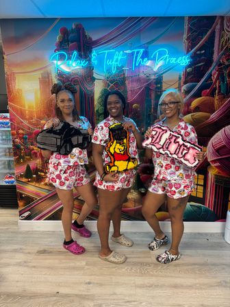 Three smiling women in matching pink patterned tops and shorts holding colorful handmade tufted rugs inside a vibrant tufting studio with a colorful mural and blue neon sign reading "Relax & Tuft The Process".
