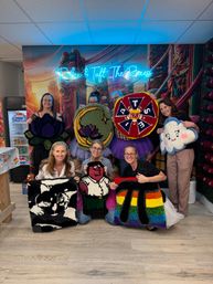 Seven women in a colorful indoor craft studio holding handmade tufted rugs—lotus, moon with flower, red-and-blue round emblem, smiling cloud, black-and-white cat, cartoon character, and rainbow pi—neon script sign and vibrant mural backdrop.