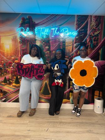 Three people in a colorful tufting studio holding handmade tufted rugs — graffiti-style 'BRATZ' piece, Sonic character rug, and a large orange flower — vibrant mural and neon sign 'Relax & Tuff The Process' in the background.