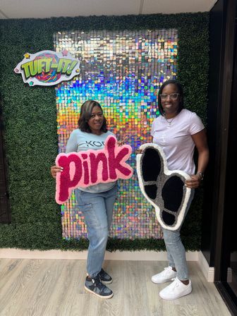 Two smiling people posing in a craft studio holding handmade tufted rugs — a bright pink "pink" word rug and a black-and-white footprint rug — against a holographic sequin and faux boxwood backdrop.