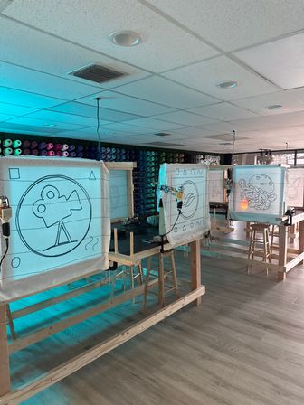 Bright indoor tufting studio with wooden frames holding stretched fabric panels sketched with designs (film camera, sea creatures), tufting guns attached, stools and workbenches, and a colorful wall of yarn spools on light wood floors.