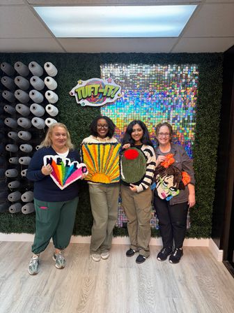 Four people in a craft studio holding colorful handmade tufted rug panels — rainbow heart, sunburst, olive oval with red dot, and a portrait — posed in front of yarn spools and a holographic backdrop