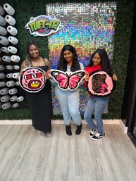 Three smiling people in a DIY tufting craft studio holding colorful handmade tufted rugs — a pink pig face, a pink butterfly, and a pink bear — posed against a holographic sequin backdrop with shelves of yarn spools.