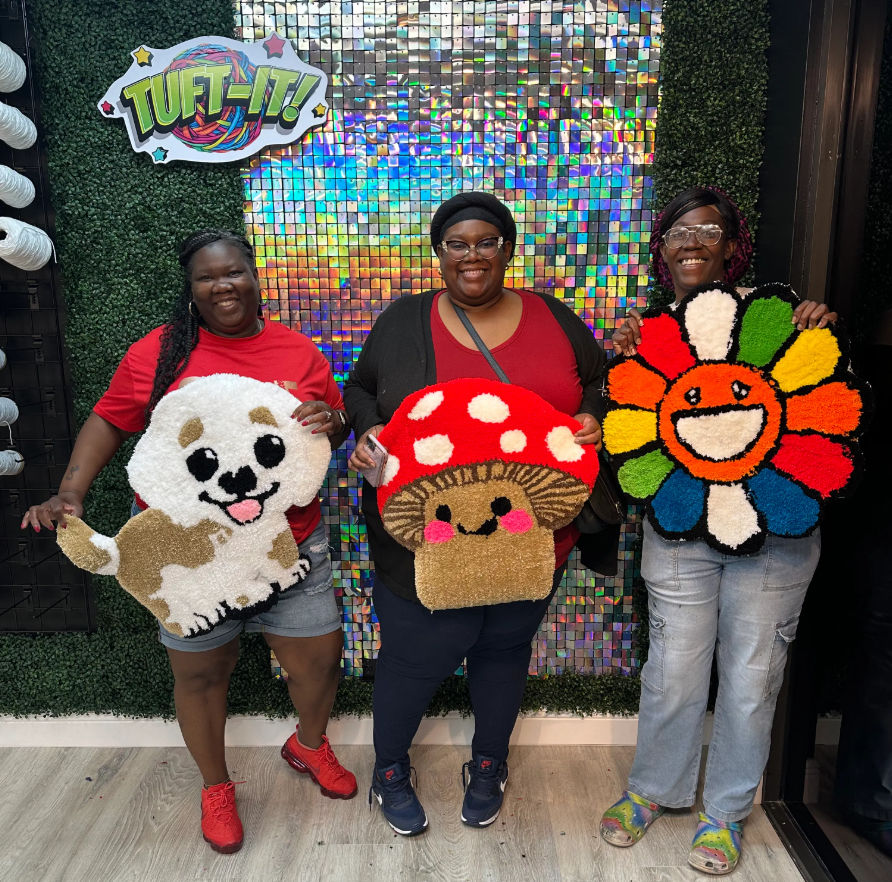Three smiling people in a tufting workshop holding colorful handmade tufted rugs — a white puppy, a red-spotted mushroom, and a rainbow smiling flower — in front of a holographic sequin backdrop.