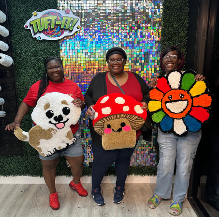 Three smiling people in a tufting workshop holding colorful handmade tufted rugs — a white puppy, a red-spotted mushroom, and a rainbow smiling flower — in front of a holographic sequin backdrop.
