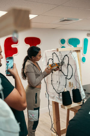 Tufting workshop scene: person using a tufting gun to stitch black yarn into a canvas on a wooden frame, forming a cartoon mouse face with yarn spools nearby and onlookers filming on a smartphone.