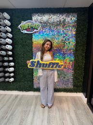Smiling woman holding a yellow-and-blue tufted “Shuffle” sign in a craft studio, posed in front of a holographic sequin backdrop framed by faux boxwood and shelves of white yarn spools.