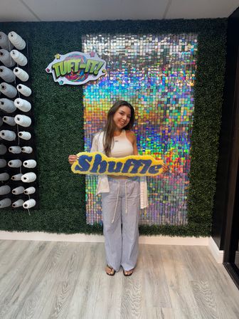 Smiling woman holding a yellow-and-blue tufted “Shuffle” sign in a craft studio, posed in front of a holographic sequin backdrop framed by faux boxwood and shelves of white yarn spools.