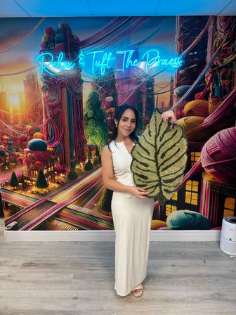 Woman in a white dress holding a large leaf-shaped tufted rug inside a colorful tufting studio with a neon sign reading "Relax & Tuft The Process" and a vibrant yarn-themed mural