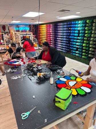 Indoor tufting studio workshop with several people making colorful tufted rugs at a large table, rainbow wall of yarn spools, tufting guns, scissors and a bright smiling flower rug in progress