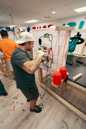 Tattooed artist in a craft studio using a tufting gun to create a cartoon dog rug on a wooden frame, with bright red yarn cones, rug scraps on the floor and colorful wall murals in the background