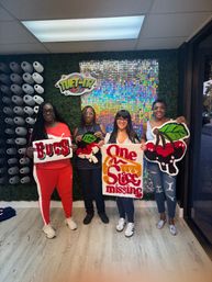 Four women smiling in a tufting workshop hold colorful handmade tufted rugs — a 'Bugs' word piece, a 'One Sock Missing' graphic rug, and cherry designs — posed against a faux boxwood wall with a sparkly sequin panel and rows of yarn spools.