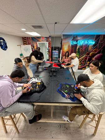 Tufting workshop in a bright craft studio: several people using tufting guns at a large table to create colorful plush rugs, with yarn, scissors, and a vivid mural on the wall.