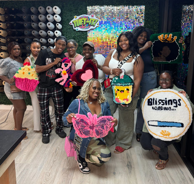Smiling group of people in a tufting workshop holding colorful handmade tufted rugs — cupcake, butterfly, heart, iced drink, crowned afro silhouette, and a round “Blessings loading” progress-bar design — posed in front of a sparkly mosaic backdrop and wall of yarn spools.