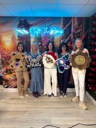 Five women at an indoor tufting workshop holding handmade tufted rugs — teddy bear, French bulldog, Santa face, floral letter 'P', and a flower with a peace sign — posed in front of a colorful mural and neon sign.