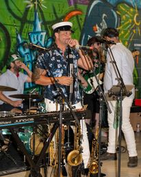 Live band performing indoors in front of a colorful graffiti mural — singer in a captain’s hat with keyboard, saxophones on stand, drums and bass guitar.