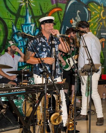 Live band performing indoors in front of a colorful graffiti mural — singer in a captain’s hat with keyboard, saxophones on stand, drums and bass guitar.