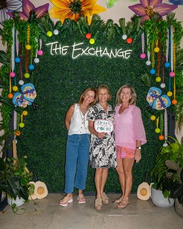 Three smiling women pose in front of a lush greenery photo wall decorated with oversized flowers, colorful hanging balls and pickleball paddles, one holding a "Queen of the Court" sign.