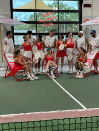 Group of people in coordinated red-and-white tennis outfits laughing and posing on an indoor court, holding paddles and sitting on striped deck chairs under a neon sign, with the net visible in the foreground.