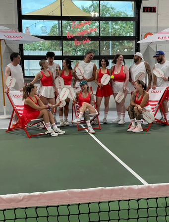 Group of people in coordinated red-and-white tennis outfits laughing and posing on an indoor court, holding paddles and sitting on striped deck chairs under a neon sign, with the net visible in the foreground.