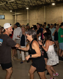 Energetic indoor warehouse party with adults dancing and mingling in casual summer clothes, many holding drinks and laughing in a crowded social gathering.