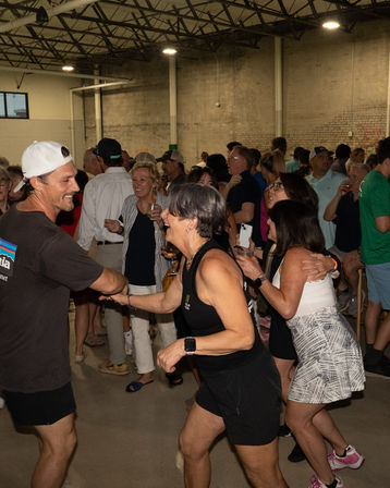 Energetic indoor warehouse party with adults dancing and mingling in casual summer clothes, many holding drinks and laughing in a crowded social gathering.