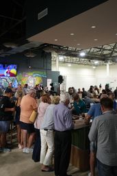 Busy indoor industrial-style bar and event space with patrons lined along a long wooden bar, bartender serving drinks, colorful graffiti mural on the wall and a large screen showing sports.