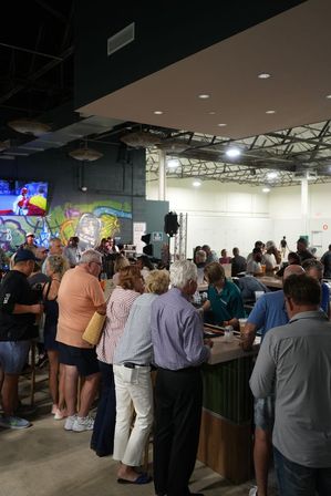 Busy indoor industrial-style bar and event space with patrons lined along a long wooden bar, bartender serving drinks, colorful graffiti mural on the wall and a large screen showing sports.
