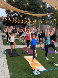 Outdoor evening yoga class on artificial turf under string lights and shade sails — a diverse group practicing single-leg balance on mats in a courtyard setting.