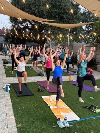 Outdoor evening yoga class on artificial turf under string lights and shade sails — a diverse group practicing single-leg balance on mats in a courtyard setting.