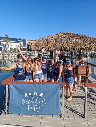 Cheering bachelorette party of women in navy tanks and captain hats on a sunny waterfront dock under a thatched tiki umbrella with a "Bachelorette Party" banner