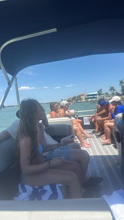 Group of people relaxing on a shaded pontoon boat on a sunny coastal bay, wearing swimsuits and caps, chatting and sipping drinks with waterfront homes and palm trees along the shoreline under clear blue skies.