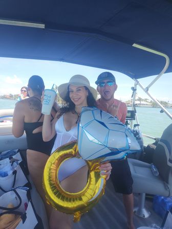 Smiling woman in a white swimsuit and sun hat holding a giant inflatable diamond ring and a drink during a sunny boat party on a coastal bay with friends in the background