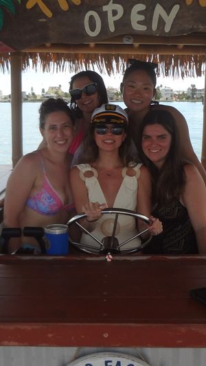 Group of five friends smiling at the helm of a floating tiki bar with a thatched roof and open sign, docked on a sunny coastal bay with an American flag and waterfront buildings in the background