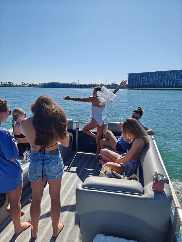 Sunny coastal scene on a pontoon boat with friends — woman in a white swimsuit and veil poses with arms out over the water while others relax on plush seating, waterfront buildings in the distance.
