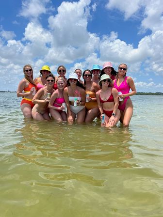 Group of women in colorful swimsuits and bucket hats standing waist-deep in shallow coastal water, holding canned drinks under a bright blue sky with puffy clouds — summer beach day by the shoreline.