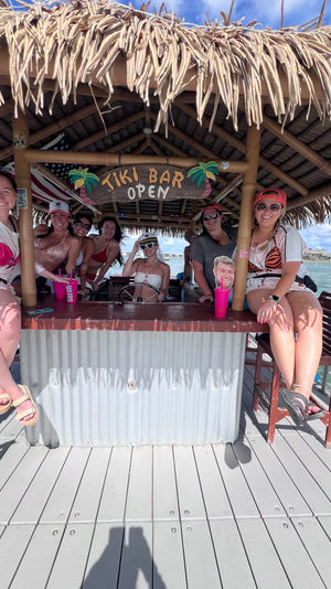 Smiling group enjoying a thatched-roof tiki bar on a floating dock boat with colorful drinks, sunny sky and coastal skyline in the background