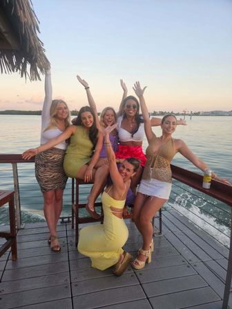 Group of six friends posing and cheering on a tropical waterfront dock at sunset, wearing colorful summer outfits and platform sandals, with ocean waves and a distant shoreline in the background.