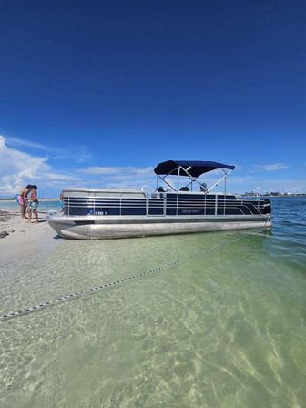 Pontoon boat beached on a white sandbar in crystal-clear shallow water under a bright blue sky, with two people standing on the shore nearby — sunny beach day.