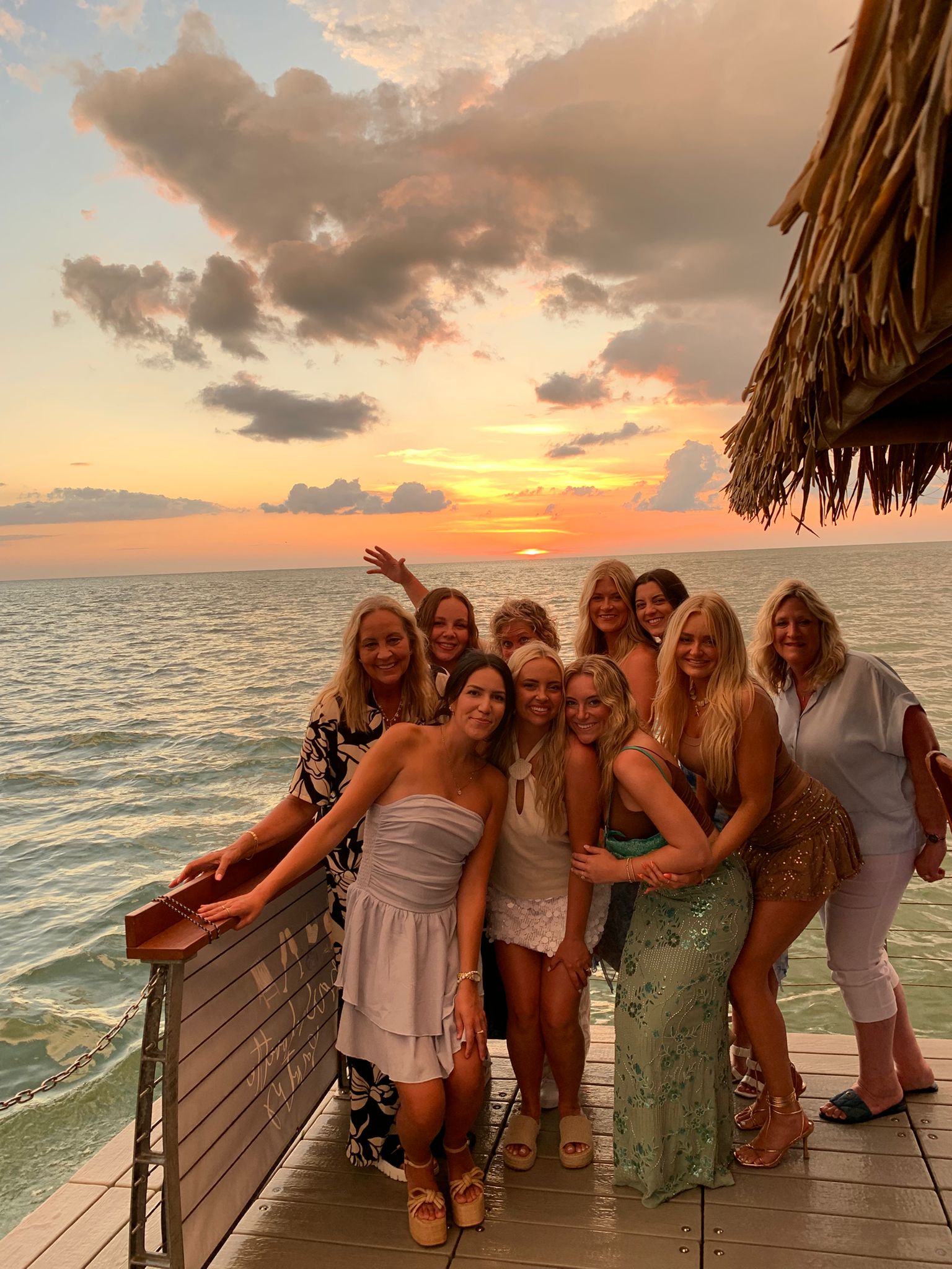 Group of smiling women posing on a wooden pier by the ocean at a tropical sunset, warm golden-pink sky with clouds and a thatched-roof edge visible — beach sunset vacation photo