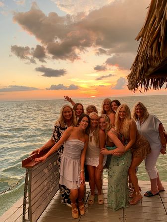 Group of smiling women posing on a wooden pier by the ocean at a tropical sunset, warm golden-pink sky with clouds and a thatched-roof edge visible — beach sunset vacation photo