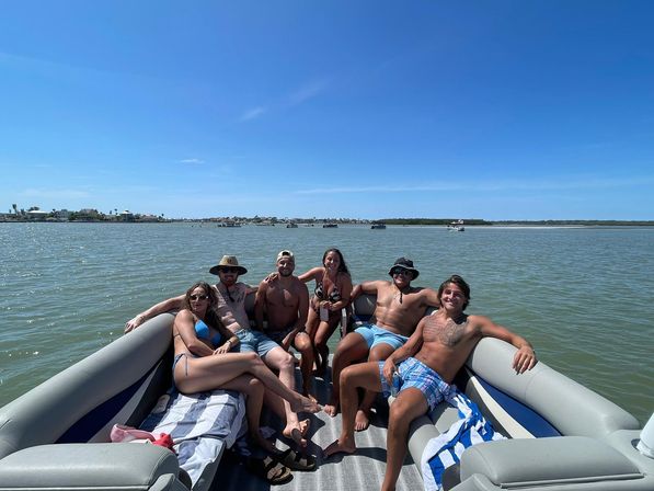 Seven people in swimsuits relaxing on a pontoon boat in a sunny coastal bay with calm water and a distant shoreline and boats