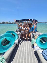 Group of friends in swimsuits cheering on a pontoon boat in turquoise coastal waters under a clear blue sky with inflatable tubes and a shoreline in the background