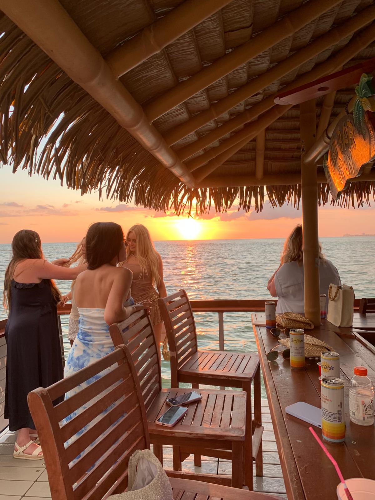 Group of people on an overwater tiki bar deck watching a vibrant tropical sunset over the ocean, thatched roof and wooden chairs with drinks and beach bags on the bar.