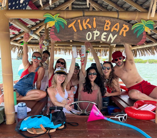 Group of friends partying on a floating tiki bar boat, raising drinks beneath a "Tiki Bar Open" sign with thatched roof, palm decor and turquoise water