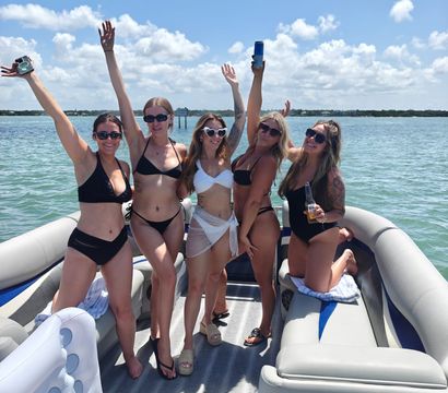 Five friends in bikinis cheering and holding drinks on a pontoon boat under a bright blue sky, turquoise coastal water and distant shoreline in the background — lively summer boating scene.