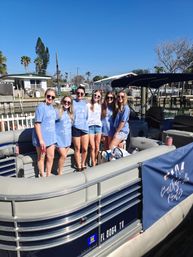 Six friends in matching light-blue 'Last Toast on the Coast' shirts smiling on a pontoon boat docked at a sunny Florida canal with palm trees and waterfront homes