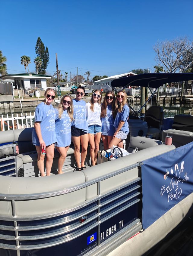 Six friends in matching light-blue shirts smiling together on a pontoon boat at a sunny Florida marina with palm trees and clear blue sky