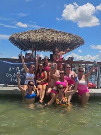 Cheerful group of women in colorful swimsuits raising drinks while sitting and wading at a lakeside dock under a thatched tiki hut on a sunny summer day — bachelorette party celebration.