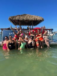 Smiling group of women in colorful swimsuits cheering with drinks at a floating tiki bar docked in shallow coastal water under a clear blue sky.